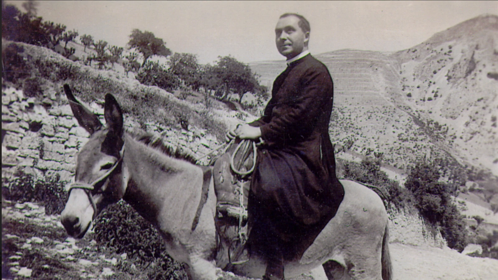 Don Achille a dorso di somaro lungo una strada di montagna a Castelletta, in una fotografia storica in bianco e nero.